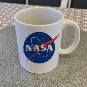May include: White ceramic mug with a blue and red NASA logo. The logo features a red curved line and a blue circle with white stars and the word "NASA" in white letters.