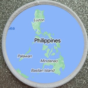 Può includere: Un cerotto circolare bianco con sfondo blu e una mappa verde delle Filippine. La mappa mostra le isole di Luzon, Palawan, Mindanao e l'isola di Basilan. Il testo "Filippine" è scritto in nero sulla mappa.