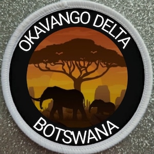 Może przedstawiać: Biała okrągła łatka z czarnym tekstem i sylwetką afrykańskiego zachodu słońca na sawannie z dwoma słoniami. Tekst brzmi "Okavango Delta Botswana".
