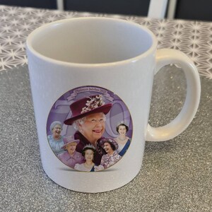 Könnte beinhalten: Weiße Keramiktasse mit einem lila und weißen Design mit einem Porträt von Königin Elisabeth II und dem Text "Platinum Jubilee 2022".