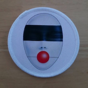 May include: White fabric patch with a black and white image of a face with a red circle for a mouth.