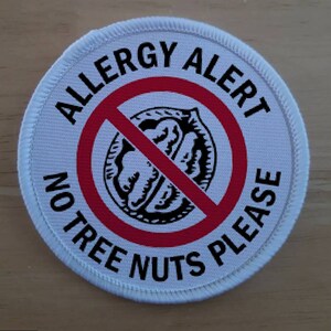 May include: White round patch with red and black text that reads "Allergy Alert No Tree Nuts Please" with a red circle and diagonal line through a black and white illustration of a nut.