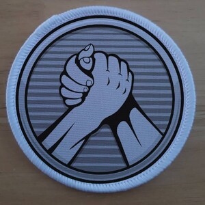 Könnte beinhalten: Ein weißer runder Patch mit einem grauen und weißen gestreiften Hintergrund. In der Mitte des Patches befindet sich ein Schwarzweißbild von zwei sich schüttelnden Händen.