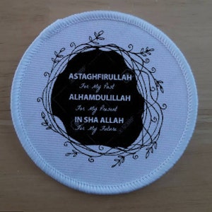Puede incluir: Un parche redondo blanco con un borde cosido. El parche presenta un diseño circular negro con el texto blanco: "ASTAGHFIRULLAH For My Past, ALHAMDULILLAH For My Present, IN SHA ALLAH For My Future". Una corona floral negra rodea el texto.