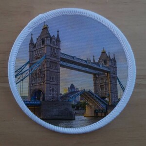 Pode incluir: Um remendo circular branco com uma foto da Tower Bridge em Londres, Inglaterra. A ponte está levantada, com o céu azul visível no fundo.
