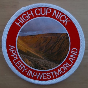 May include: A white round patch with a red border. The patch has a picture of a valley with a mountain in the background. The text "HIGH CUP NICK APPLEBY-IN-WESTMORLAND" is written in a circle around the picture.