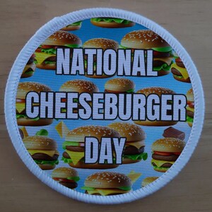 May include: White round patch with a blue background and a white border. The patch has a pattern of cartoon cheeseburgers and the text "NATIONAL CHEESEBURGER DAY".