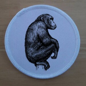 May include: A white round patch featuring a black and white illustration of a chimpanzee sitting on a branch. The chimpanzee is facing to the right and has a thoughtful expression.