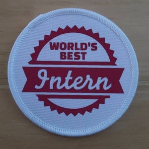 Könnte beinhalten: Runder weißer Patch mit roten Nähten am Rand. Der Patch hat ein rotes Sternburst-Design mit dem Text "WORLD'S BEST Intern" in Weiß.