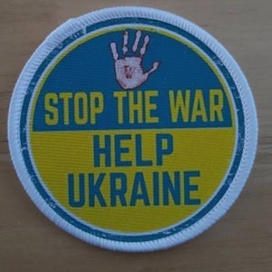May include: A round, white fabric patch with a blue and yellow circle in the center. The circle has a white handprint in the top half and the text "STOP THE WAR HELP UKRAINE" in the bottom half.