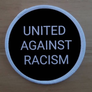 Può includere: Un cerchio bianco con un fondo nero e il testo "UNITED AGAINST RACISM" in bianco.