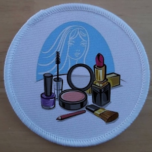 Peut inclure: Un patch rond blanc avec un fond bleu et une silhouette blanche d'une femme aux cheveux longs. Le patch présente une variété d'articles de maquillage, notamment un rouge à lèvres, un vernis à ongles, un fard à paupières et un pinceau à maquillage.