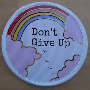Könnte beinhalten: Runder weißer Patch mit einem Regenbogen, Wolken und dem Text "Don't Give Up".