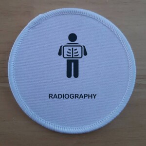 May include: White round fabric patch with black stitching around the edge. The patch features a black silhouette of a person holding an x-ray image of a skeleton. The text "RADIOGRAPHY" is printed in black below the image.