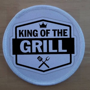 Könnte beinhalten: Weißer runder Patch mit schwarzem Text und einer Krone mit der Aufschrift "King of the Grill" und darunter ein überkreuztes Spatel und eine Gabel.