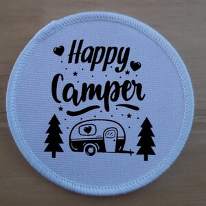 May include: White round fabric patch with black text that says "Happy Camper" and a black illustration of a camper, two pine trees, and stars.