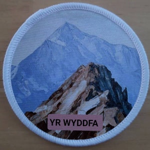 Puede incluir: Un parche redondo blanco con una escena de montaña azul y blanca. El texto "YR WYDDFA" está impreso en un rectángulo rosa en la parte inferior del parche.