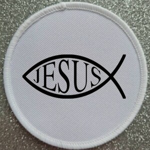 Op de afbeelding: Ronde witte stoffen patch met een zwarte omtrek van een vis met het woord "JESUS" erin.