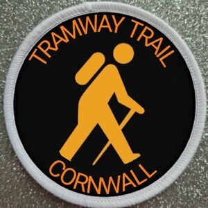 Pode incluir: Um remendo circular branco e preto com o texto "Tramway Trail Cornwall" em laranja. Um caminhante estilizado laranja com uma mochila e um bastão de caminhada está no centro do remendo.