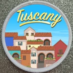 Könnte beinhalten: Ein runder, weißer Patch mit blauem Hintergrund und dem Wort "Toskana" in gelber Schrift. Der Patch zeigt eine Cartoon-Illustration einer Reihe von bunten Häusern mit roten Ziegeldächern.