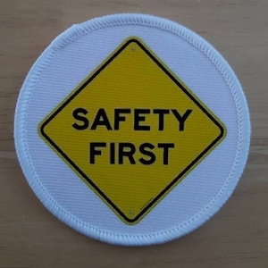 May include: A white round patch with a yellow diamond-shaped sign that says "SAFETY FIRST" in black letters.