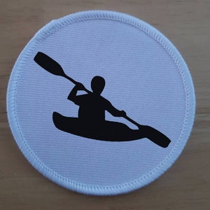 Pode incluir: Etiqueta redonda de tecido branco com uma silhueta preta de uma pessoa em caiaque. A pessoa está remando com um único remo.