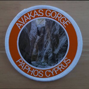 May include: A white round patch with an orange border. The patch has a picture of a narrow canyon with the text "AVAKAS GORGE PAPHOS CYPRUS" around the image.