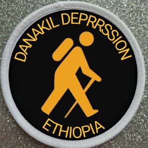 May include: A black and white circular patch with a white border. The centre of the patch features a yellow stick figure hiking with a backpack and a walking stick. The text "DANAKIL DEPRESSION ETHIOPIA" is written in yellow around the stick figure.