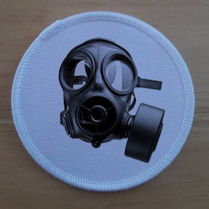 Op de afbeelding: Ronde patch met een gedetailleerd zwart-grijs gasmasker ontwerp. Het gasmasker staat centraal op een lichtblauwe achtergrond, met een wit gestikte rand. Geschikt voor kleding of accessoires.