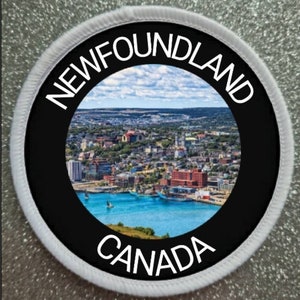 May include: A round, white patch with a black circle in the center. The circle has the words "NEWFOUNDLAND CANADA" in white text around a photo of a city skyline and a body of water.