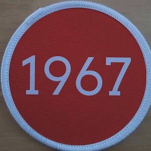 Puede incluir: Un parche circular rojo con un borde blanco y el número 1967 impreso en blanco.