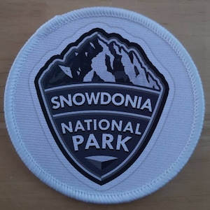 Może przedstawiać: Biała okrągła łatka z szarym logo w kształcie tarczy. Logo przedstawia pasmo górskie i tekst "Snowdonia National Park".