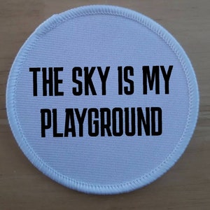 Può includere: Toppa rotonda in tessuto bianco con testo nero che recita "The sky is my playground".