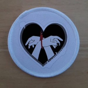 May include: A white fabric patch with a black heart shape containing a graphic of two hands tied together with a red rope.