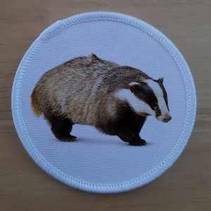 May include: A white fabric patch with a brown and white badger printed on it. The patch has a white border with a stitched edge.