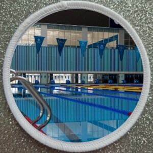 May include: A view of a swimming pool through a round window. The pool is blue and has multiple lanes marked with black lines. The pool is surrounded by a white tiled wall and a white ceiling.