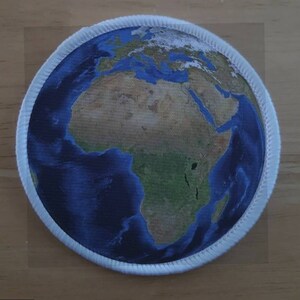 May include: A white circular patch with a blue and green image of the Earth, centred on Africa. The patch has a white stitched border.