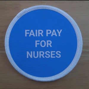 May include: A blue round fabric patch with white stitching around the edge. The text "FAIR PAY FOR NURSES" is printed in white on the patch.