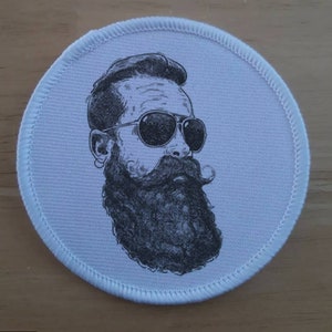 以下が含まれることがあります： 白い丸いパッチに、黒と白のイラストで描かれた、ひげを生やしたサングラスをかけた男性。男性は口ひげとトップノットをしています。