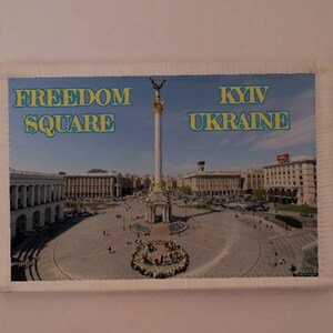 May include: A postcard featuring a photo of Freedom Square in Kyiv, Ukraine. The square is surrounded by buildings and a monument with a statue on top. The text "FREEDOM SQUARE" and "KYIV UKRAINE" is printed on the postcard.