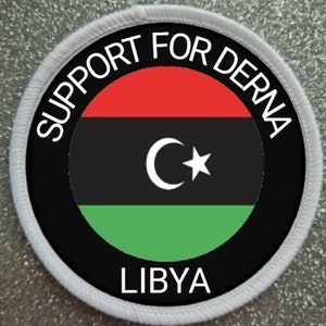 May include: A white circular patch with black and white text that reads "Support for Derna Libya". The centre of the patch features the Libyan flag, which is a horizontal triband of red, black, and green with a white crescent moon and star.