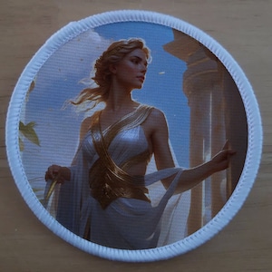 May include: A round white patch with a blue background featuring a woman with long blonde hair wearing a white dress with gold accents. The woman is standing in front of a white pillar with a blue sky in the background.