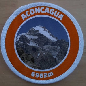 Puede incluir: Parche redondo con la palabra "ACONCAGUA" en blanco sobre un borde naranja. El centro presenta una escena de montaña con picos nevados sobre un cielo azul. La parte inferior del parche dice "6962m", que son 6.962 metros.