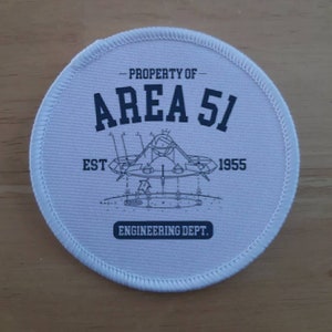 May include: White round patch with black text and a black and white illustration of a flying saucer. The text reads "PROPERTY OF AREA 51 EST 1955 ENGINEERING DEPT."