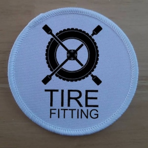Pode incluir: Um patch redondo branco com um design preto de pneu e chave de porcas. As palavras "TIRE FITTING" são impressas em preto abaixo. O patch tem uma borda branca costurada.