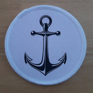 Peut inclure: Un patch rond blanc avec un motif d'ancre noire. L'ancre a un anneau en haut et deux bras avec des boules rondes aux extrémités.