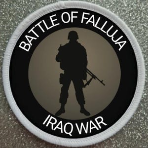 May include: A circular patch with the text "BATTLE OF FALLUJA" and "IRAQ WAR" in white. A black silhouette of a soldier holding a rifle is in the center, set against a gray background within a black circle.