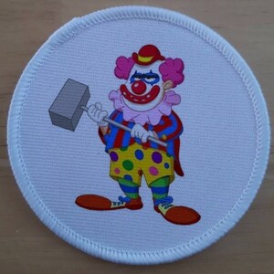 May include: Round white patch featuring a cartoon clown with pink hair, a red nose, and a grumpy expression. The clown is holding a gray mallet. The clown wears a red hat, a ruffled collar, and a colorful striped jacket and polka dot shorts.