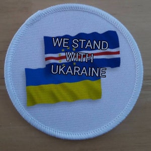 May include: A white round patch with the text "WE STAND WITH UKRAINE" and the flags of Ukraine and Cape Verde.