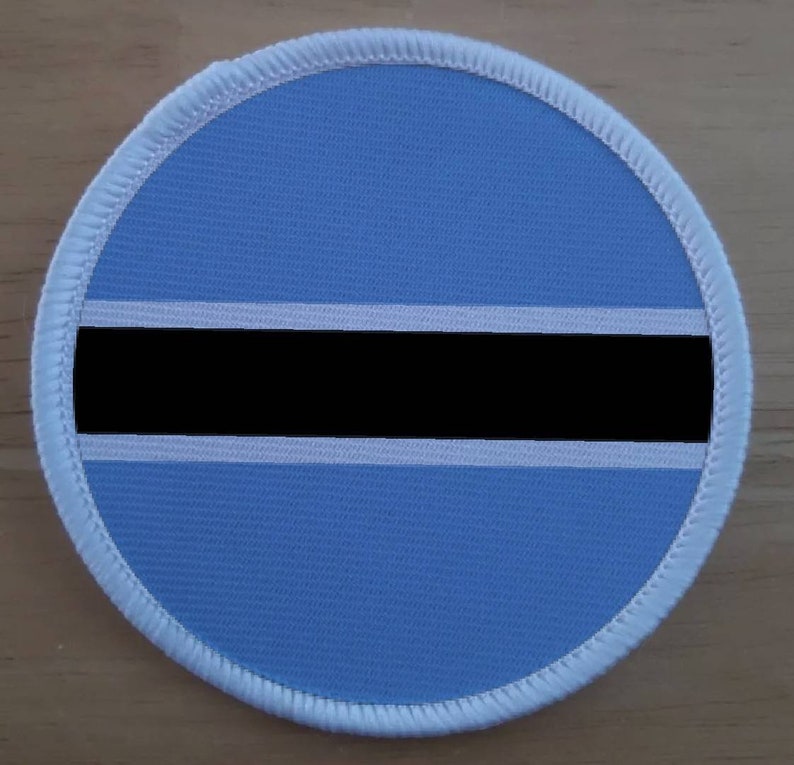 Pu&ograve; includere: Un cerotto rotondo in tessuto blu con un bordo bianco. Il cerotto presenta una striscia orizzontale nera con due strisce bianche sopra e sotto.